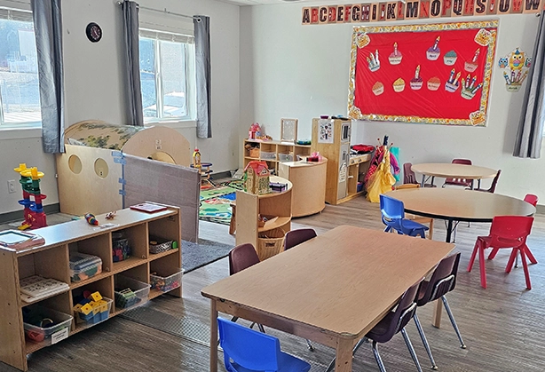 Preschool classroom with small tables, colorful chairs, educational toys, and a play area, featuring bright natural light and a birthday-themed bulletin board